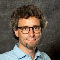 Portrait von Dustin Wiemann, Mann mit kurzen Locken, etwas Bart, Brille und einem hellblauen Hemd.