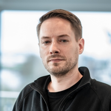 Portraitfoto von Simon Frerichs, Leiter IT-Infrastruktur bei LWLcom, trägt ein schwarzes T-Shirt und eine schwarze Jacke mit Reißverschluss, steht vor einem unscharfen Hintergrund.